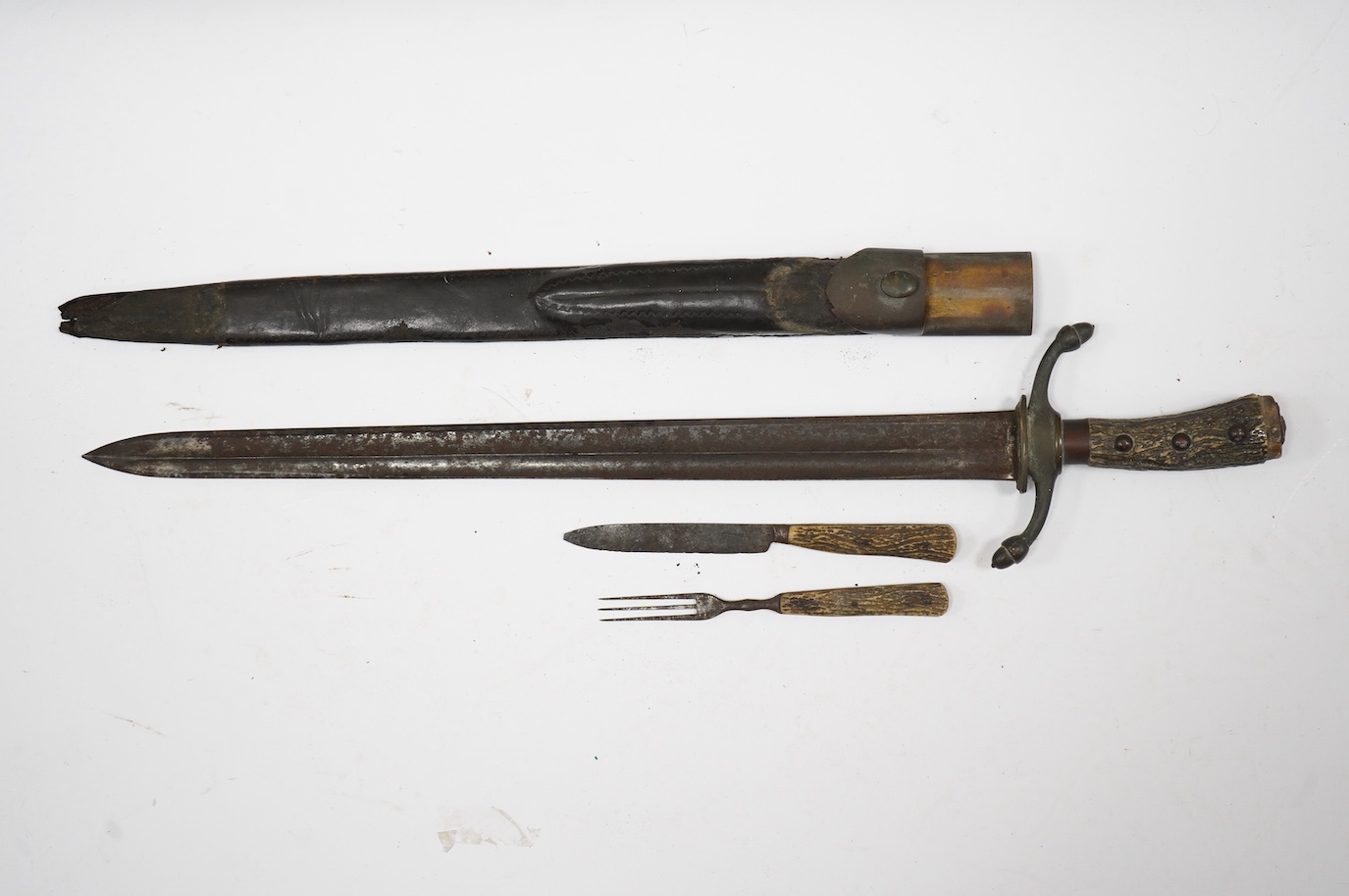 A 19th century Continental hunting hanger, blade cut with twin fullers, recurved brass crosspiece with acorn finials, staghorn grip (pommel missing), in its brass mounted leather scabbard, containing knife and fork (chap